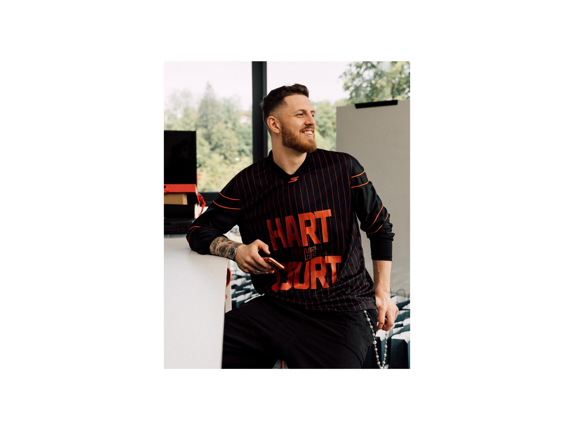 Portrait of a man smiling in a black and orange HART OF THE COURT jersey by a window